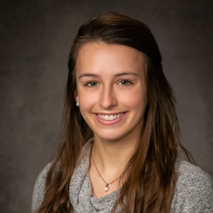 Female with long brown wavy hair pinned back wearing a gray V-neck sweater